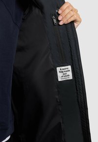 Schwarze gepolsterte Jacke mit Innenfutter, einem Reißverschlussfach und einem Pflegeetikett, das besagt: "Eine warme Umarmung aus Stoff." Hat eine glatte Textur.