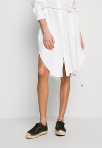 Rochie albă din cămașă cu talie încinsă, având mâneci lungi și fante laterale. Pur și simplu cu pantofi espadrile negri cu șireturi și talpă din jută.