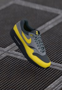 Nike Air Max 1 sneaker featuring grey and yellow paneling, textured mesh and suede materials, with black accents and laces.