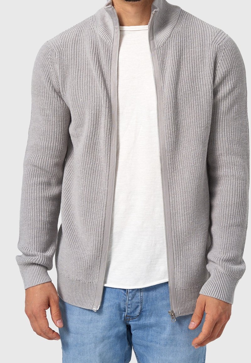Homme portant un pull zippé côtelé gris clair par-dessus un t-shirt blanc et un jean bleu, debout les mains le long du corps devant un fond neutre.