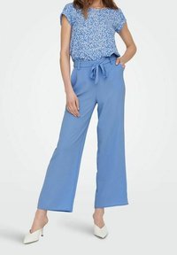 Donna che indossa una blusa corta a maniche corte blu con fiori, infilata in un pantalone ampio a vita alta blu con cintura legata, abbinato a tacchi bianchi.