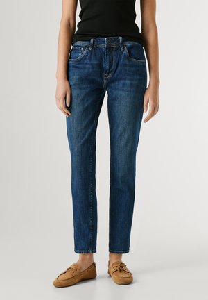 Personne portant un jean droit bleu, un haut sans manches noir et des mocassins en daim marron, debout devant un fond uni.