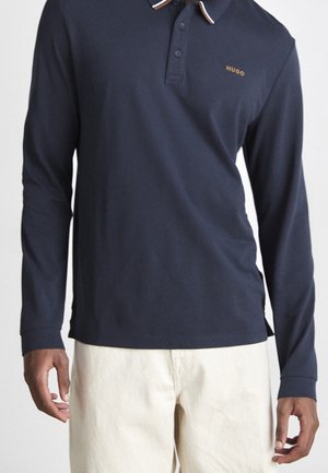 Hombre con polo de manga larga azul marino con ribete blanco y naranja en el cuello y pantalones beige, de pie con los brazos relajados.