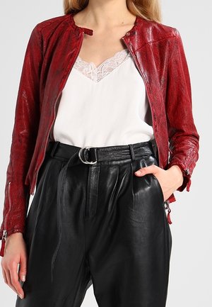 Femme portant une veste en cuir rouge texturée, un caraco blanc orné de dentelle, et un pantalon en cuir noir taille haute avec une ceinture à double anneau.