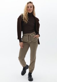 Maglione a collo alto marrone a coste, caldo cardigan marrone, pantaloni jogger verde oliva con polsini elasticizzati e stivali neri con lacci. Outfit informale con dettaglio a cordoncino.