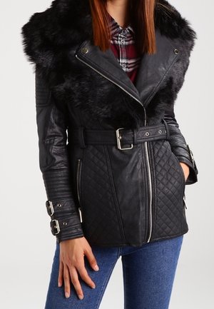 Femme portant une veste matelassée en cuir noir avec col en fourrure, taille ceinturée, poches zippées et poignets à boucle, sur une chemise à carreaux rouge et un jean bleu.