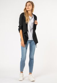 Zwarte lichte jas met een open voorkant, opgerolde mouwen en een trekkoordzoom, gecombineerd met een effen wit T-shirt en lichtblauwe jeans.