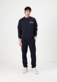 Marineblå sweatshirt og matchende joggers i blødt stof. Sweatshirten har et hvidt logo på brystet. Modellen har hvide sneakers på.