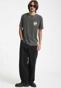Mørkegrå t-shirt med et rundt hvidt grafik på venstre bryst, kombineret med løse sorte bukser og sorte sko, med en glat tekstur.