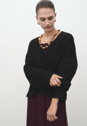 Cardigan nero lavorato a maglia con scollo a V e chiusura con bottoni, con polsini a coste. Indossato sopra un abito bordeaux, accessoriato con una collana multicolore.