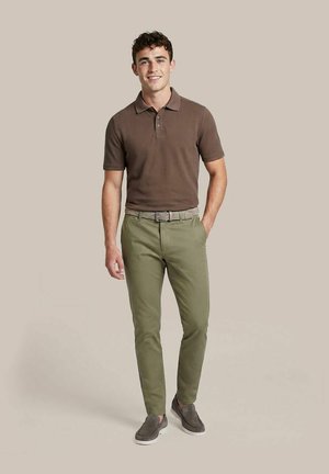 Jonge man die lacht, draagt een bruin poloshirt, groene broek met een gevlochten riem en grijze instapschoenen, staat met één hand in de zak.