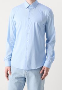 Camisa de manga larga color azul claro confeccionada en tela lisa. Cuenta con cuello puntiagudo, cierre de botones y un logo sutil en el pecho izquierdo.