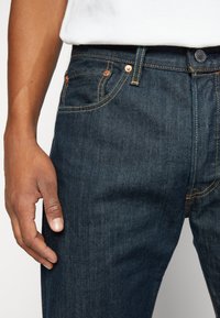 Vaqueros de mezclilla azul oscuro con corte slim, con detalles de cobre en los bolsillos y una costura visible a lo largo de las costuras.