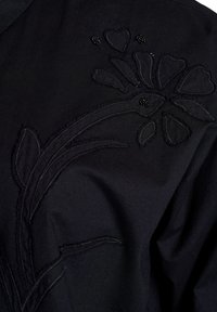 Zwarte stoffen top met bloemen borduursels in textuur van zwart garen en met kralen accenten, met nadruk op hartvormen en bladeren.