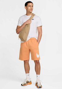 Pantalones cortos de sudadera naranjas con un logo blanco, camiseta blanca, bandolera marrón claro y zapatillas en tonos neutros con detalles texturizados.