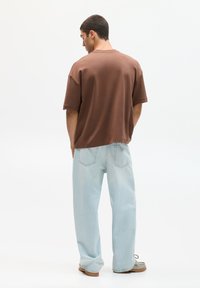 Hnědé oversized tričko s krátkými rukávy, uvolněný střih. V kombinaci se světle modrými džíny, viditelné švy a hnědé obuv.