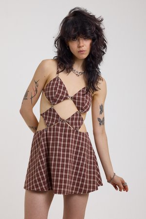 Kvinne med mørkt bølget hår iført en brun rutete kjole med utskjæringer, som viser tatoveringer på armene, og sølvsmykker mot en ensartet bakgrunn.