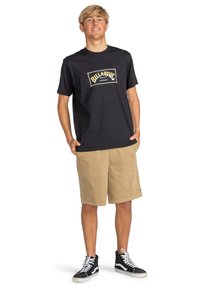 Black cotton t-shirt with yellow and white logo on chest, paired with beige corduroy shorts and black high-top sneakers.