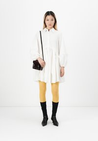 Kvinne iført en løs hvit kjole med ballongsleeves, sennepgule leggings, svarte sokker, svarte spisse sko og en liten mørk crossover-veske.
