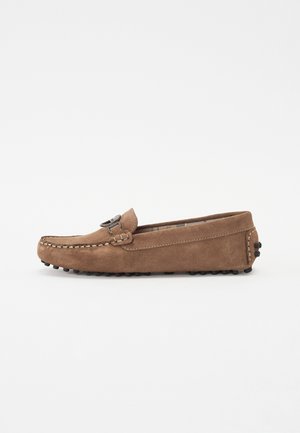 Mocassin en daim marron clair sans lacets avec une boucle métallique, coutures visibles et picots en caoutchouc sur la semelle et le talon.