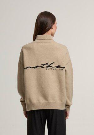Beige oversized sweater med høj krave, prydet med sort broderet tekst "mother COTTON LAB" på ryggen. Blød, strikket tekstur.