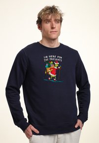 Sudadera de color azul marino con un gráfico impreso de un personaje vestido con ropa navideña, esquiando con una bolsa de regalos. Puños y dobladillo acanalados.