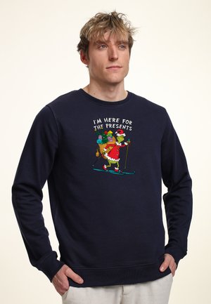 Marinblå tröja med tryckt grafik av en karaktär i julaftonskläder som åker skidor med en säck av presenter. Ribbad mudd och nederkant.