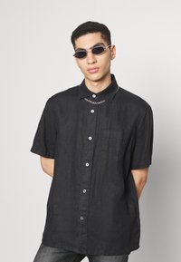 Camisa preta de manga curta com botões e bolso, feita de tecido texturizado. Colar de corrente prateada e óculos de sol redondos completam o visual.