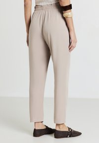 Persona che indossa pantaloni elasticizzati beige con vita alta, scarpe slip-on in tessuto scuro e due braccialetti larghi sul braccio sinistro, in piedi su un pavimento chiaro.