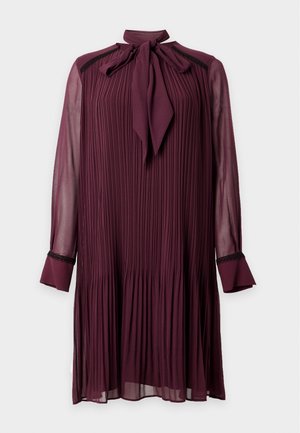 Un mannequin porte une robe plissée bordeaux jusqu’aux genoux, avec un col à nœud, et des bottines noires. Il se tient devant un fond blanc uni de studio.