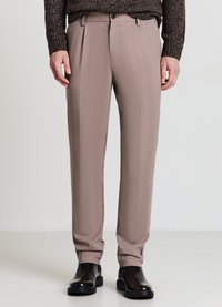 Hellbraune, maßgeschneiderte Hose mit glatter Textur, ausgestattet mit einer vorderen Plissee, Gürtelschlaufen und einem einzelnen Knopfverschluss. Kombiniert mit schwarzen Stiefeln.