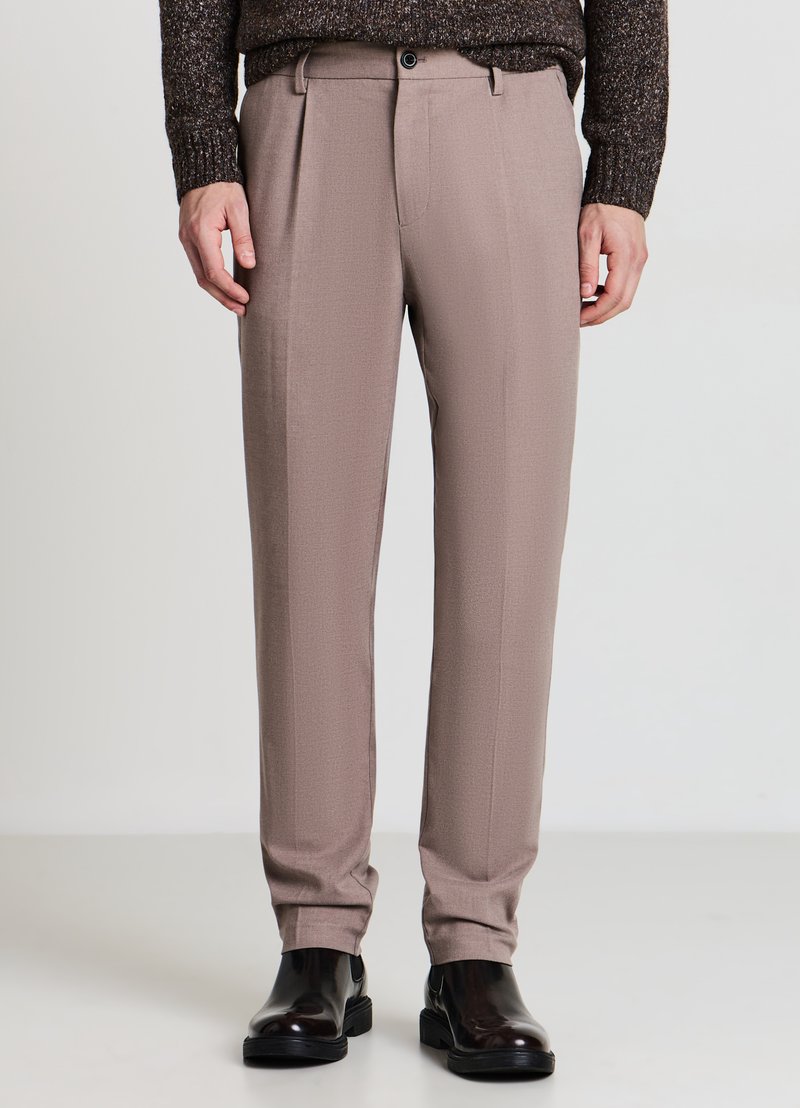 Hellbraune, maßgeschneiderte Hose mit glatter Textur, ausgestattet mit einer vorderen Plissee, Gürtelschlaufen und einem einzelnen Knopfverschluss. Kombiniert mit schwarzen Stiefeln.