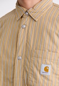 Tan striped shirt made from soft fabric, featuring a buttoned collar, silver buttons, and a logo patch on the chest pocket.