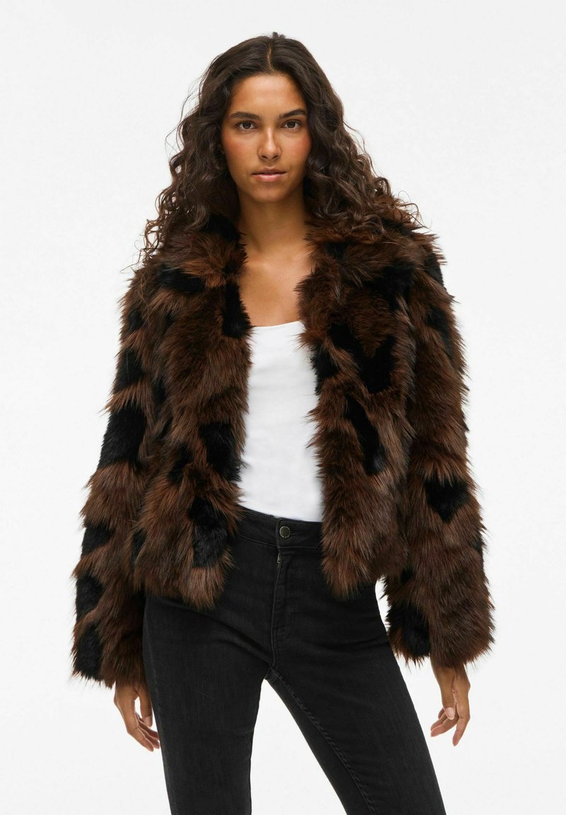 Mākslīgā kažokāda ar brūna un melna laukumu dizainu, ar apkakli un garām piedurknēm. Uzvilkta virs baltas blūzes un melnām džinsām.