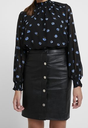 Kvinde iført en sort bluse med lange ærmer med blå blomsterprint, stukket ned i et sort lædernederdel med knapper foran.