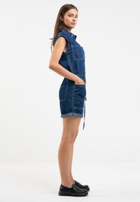 Tamsiai mėlynas džinsinis kombinezonas su trumpomis ranksovėmis. Yra dvi priekinės kišenės, sukdami rankogaliai ir diržas juosmeniui reguliuojamam pritaikymui. Lygus paviršius.