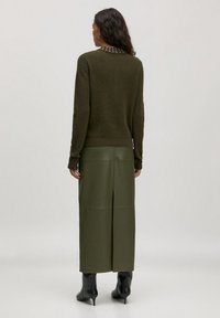 Vrouw met lang donker haar, gekleed in een olijfgroene trui en een bijpassende lange leren rok met een split aan de achterkant, en zwarte leren enkellaarsjes, staand.