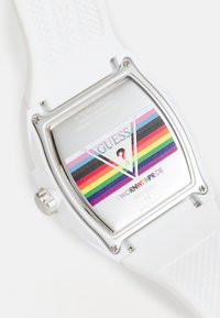 Witte rechthoekige horloge met een gestructureerde siliconen strap. De wijzerplaat heeft regenboogstrepen en het "GUESS"-logo. Waterbestendig, gepolijste stalen achterkant.