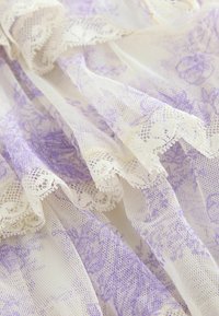 Tessuto in pizzo con motivo floreale lavanda su sfondo bianco trasparente, bordi smerlati e una texture delicata.