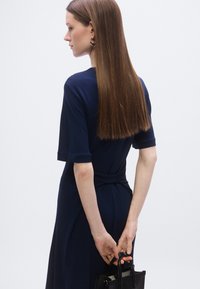 Rochie bleumarin cu mâneci scurte, detaliu cu cordon în talie, textură netedă și siluetă clasică; asortată cu o geantă mică neagră.