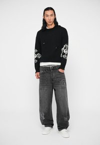 Pull en tricot noir avec un design de tête de mort blanche sur les manches, associé à un jean en denim gris ample avec des accents imprimés, et des baskets blanches.