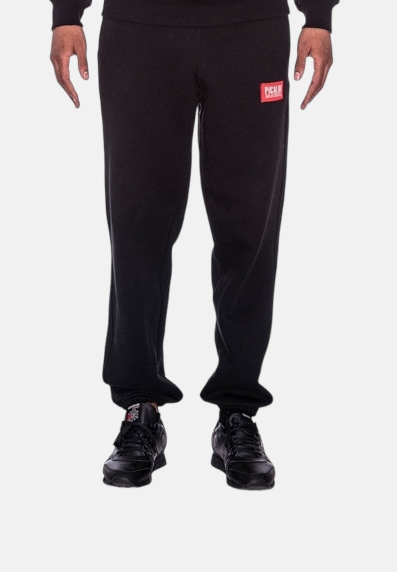 Schwarze Sweatpants aus weicher Stoff, mit einem roten Logopatch auf der linken Seite, in lockerer Passform und elastischen Bündchen an den Knöcheln.