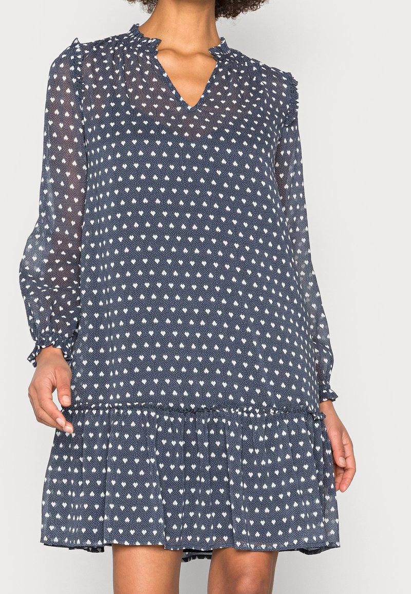 Robe bleu marine à manches longues avec imprimé losanges blancs, col à volants, poignets froncés et ourlet volanté portée par une personne visible du cou aux genoux.