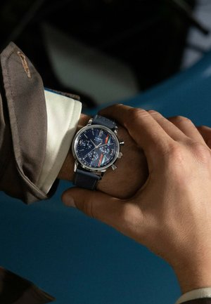 Montre chronographe avec un cadran bleu et des accents argentés, boîtier en métal, bracelet en cuir bleu marine, trois sous-cadrans et chiffres arabes.