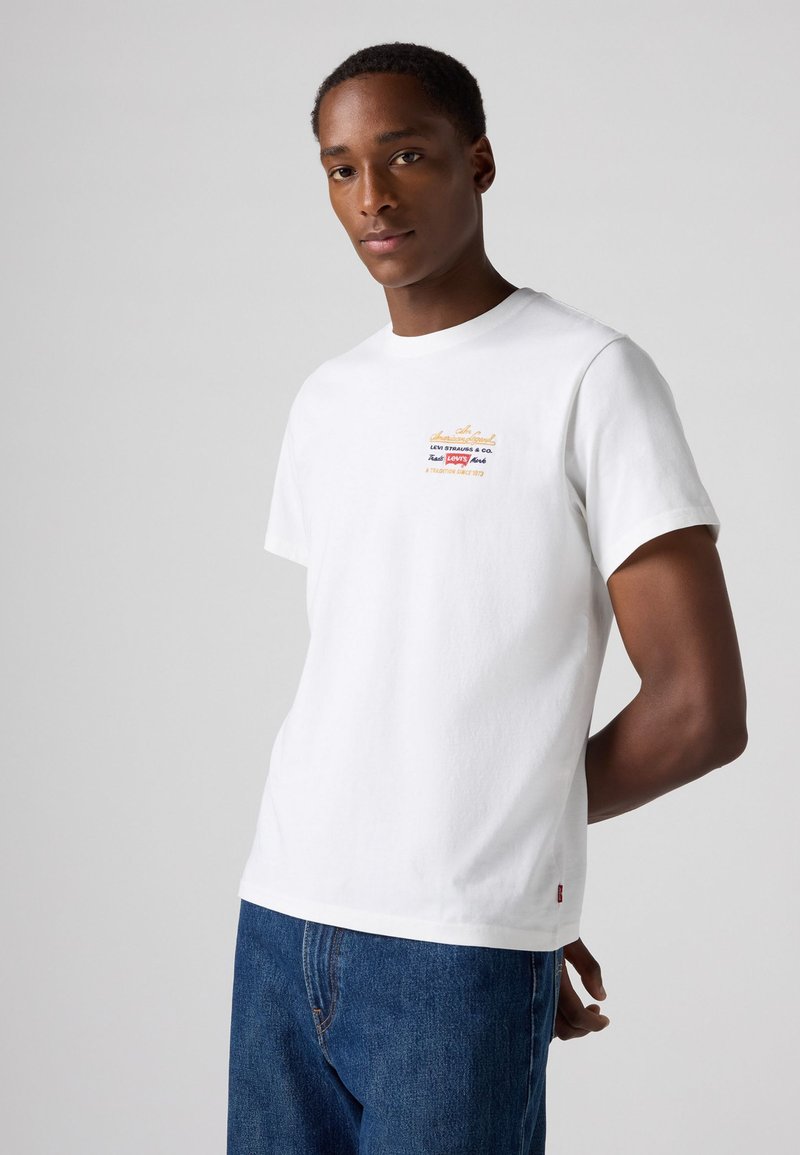 Witte katoenen t-shirt met een kleurrijk logo op de borst, ronde halslijn en korte mouwen, gecombineerd met blauwe spijkerbroek.