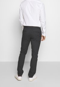 Muž v tmavě šedých na míru šitých kalhotách, bílé košili a bílých teniskách, stojící čelem od diváka na jednobarevném světlém pozadí.