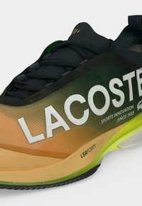Κοντινή λήψη αθλητικού παπουτσιού Lacoste με μαύρα κορδόνια, μαύρο και πορτοκαλί φλογερό επάνω μέρος, λευκό λογότυπο και ανοιχτοπράσινη σόλα με την ένδειξη LSRFOAM.