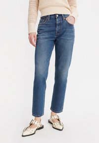 Jeans de denim azul claro con corte recto, cintura alta y ligero desgaste. Combinados con un suéter de punto color crema y zapatos crema con tachuelas.