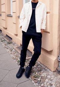 Chaqueta de algodón blanca sobre una camiseta negra, combinada con pantalones de chándal negros y zapatillas negras. El entorno presenta paredes beige y pavimento de adoquines.