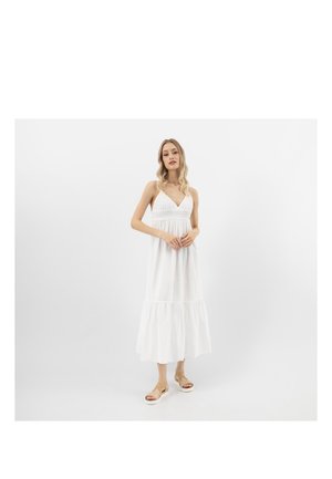 Donna che indossa un vestito midi bianco senza maniche con un orlo arricciato e sandali beige con zeppa, in piedi davanti a uno sfondo bianco uniforme.